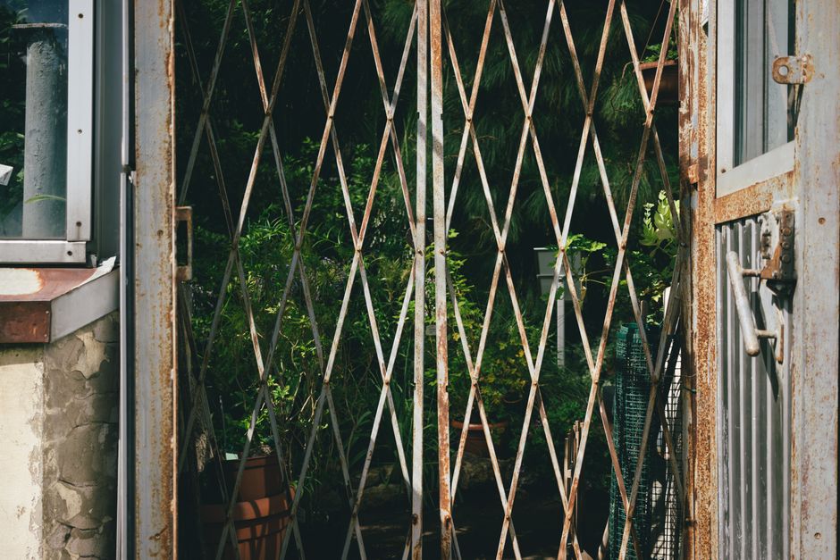 A rusted metal garden gate with a diamond-shaped lattice pattern partially open, revealing a lush, well-maintained garden behind. The garden features a variety of green plants and shrubs, some in terr